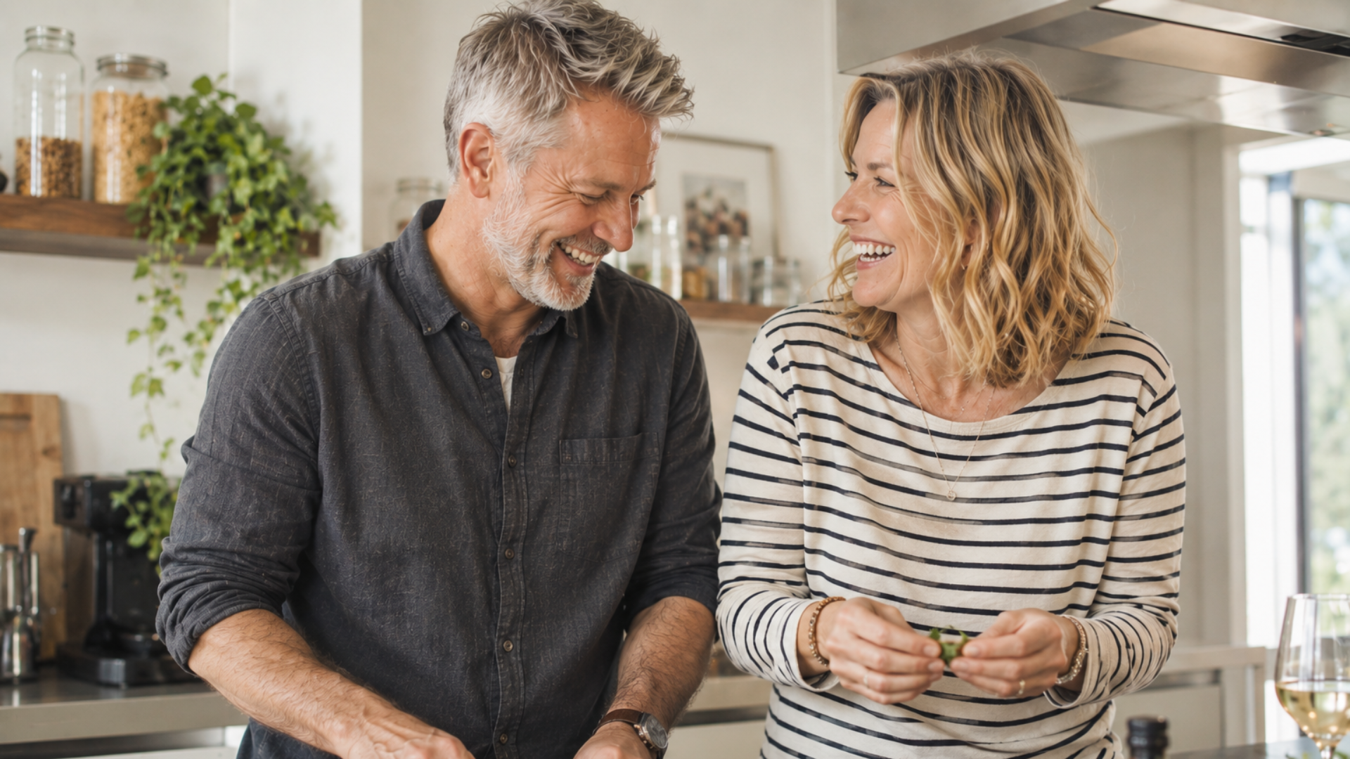 A stylish couple in their sixties laugh together while preparing dinner in a bright, modern kitchen, surrounded by fresh ingredients and greenery.